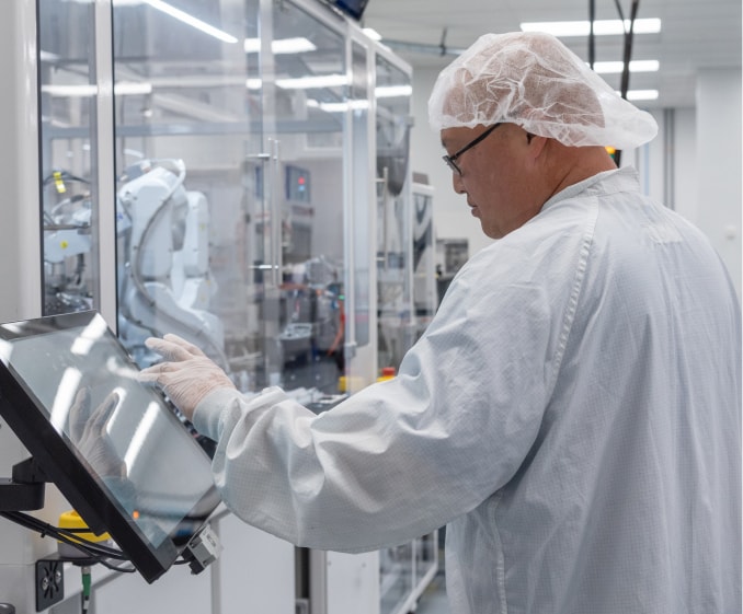 Photo of an Isometric Micro Molding employee in white coat and hair net, in Class 7 clean room, next to micro automated assembly equipment.