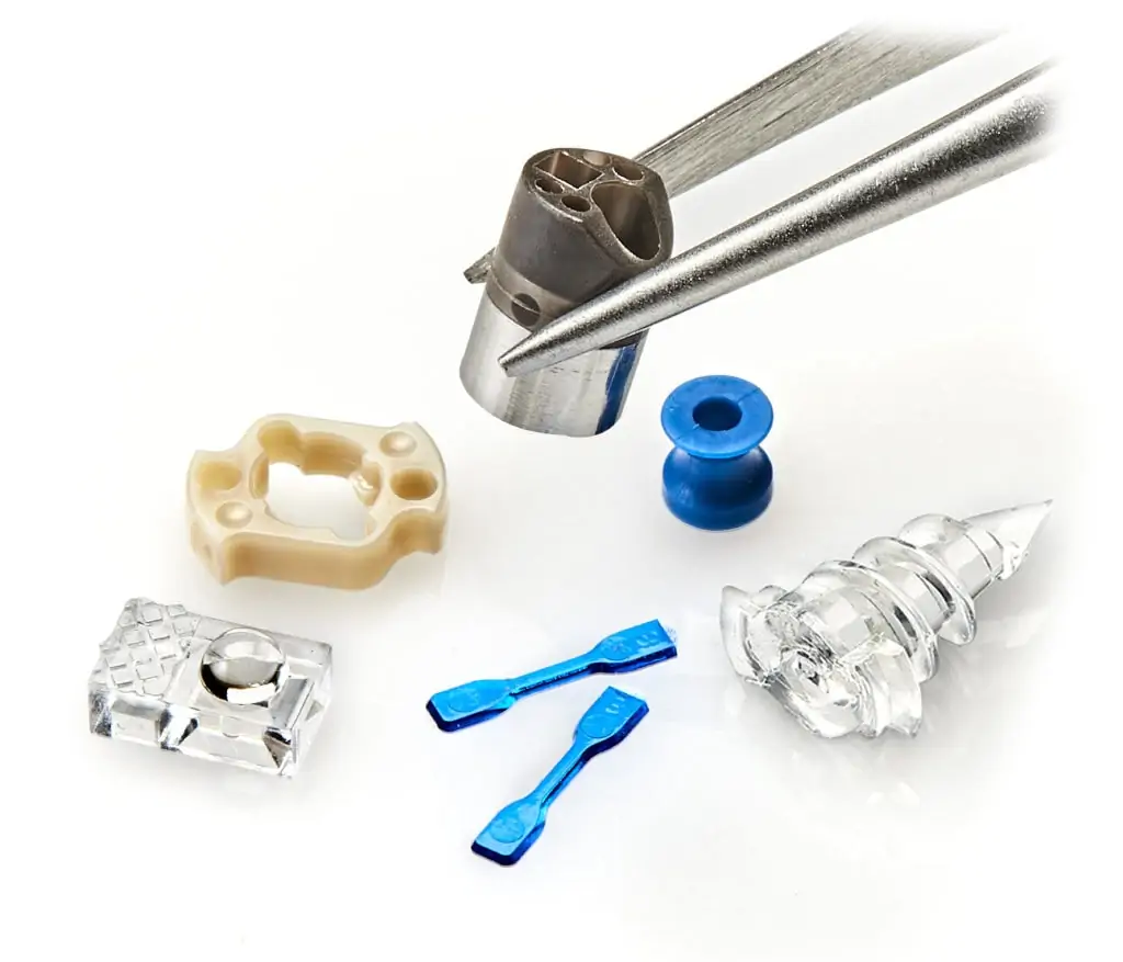 Photo of micro components including optical lens, ear drain tube, tissue repair, catheter tip, shown against a tweezer, by Isometric Micro Molding