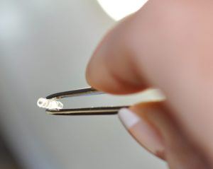 Picture of opthalmic intraocular drug delivery implant device, held by tweezer tips, by Isometric Micro Molding