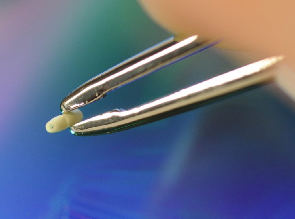 Photo of orthopedic suture anchor medical device held by tweezer tips, with thin wall molding and micro features, by Isometric Micro Molding