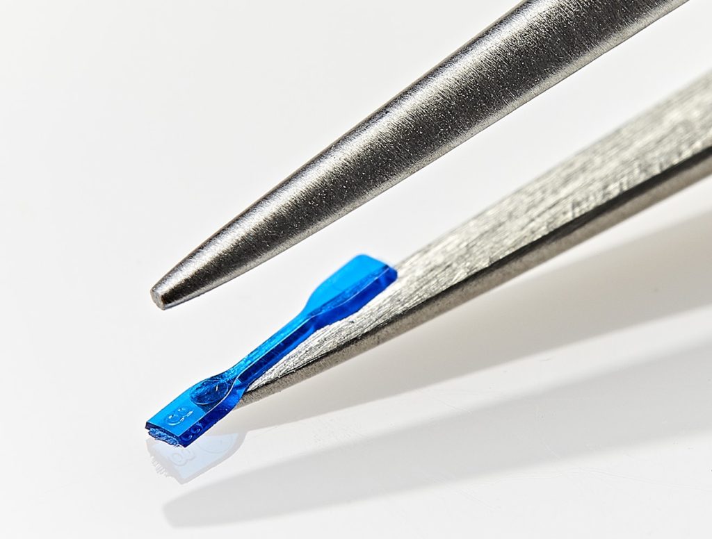 Photo of Isometric Micro Molding's micro injection molding resin flow evaluation test bar, shown on the tip of a tweezer.
